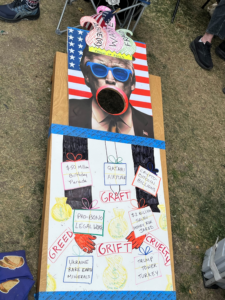 Corn hole board from the Indivisible Denver Action booth at the Denver No Kings Rally.  The hole in the board is Donald Trump's mouth and the board shows grievances such  as greed and grift.