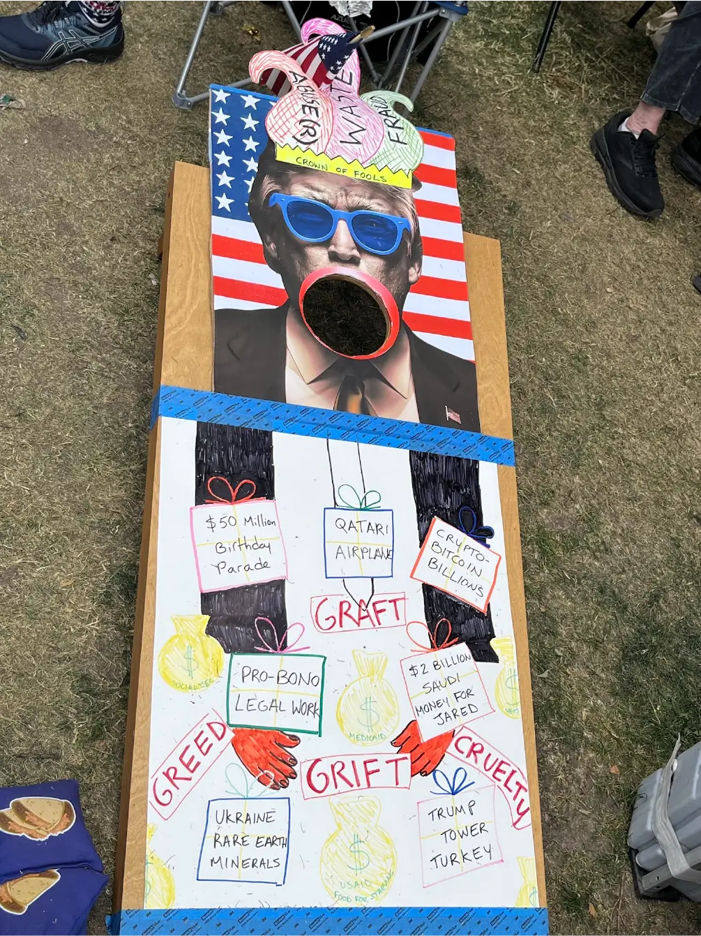 ourstory_img3webp unflattering image of Donald Trump with things that he should be ashamed of written on the sign like '$50 Million Birthday Parade'