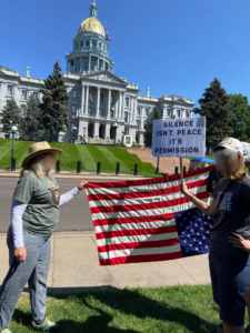 People holding an upside down flag and a sign that says, "Silence isn't Peace it's Permission."  The Colorado capital building is in the background.